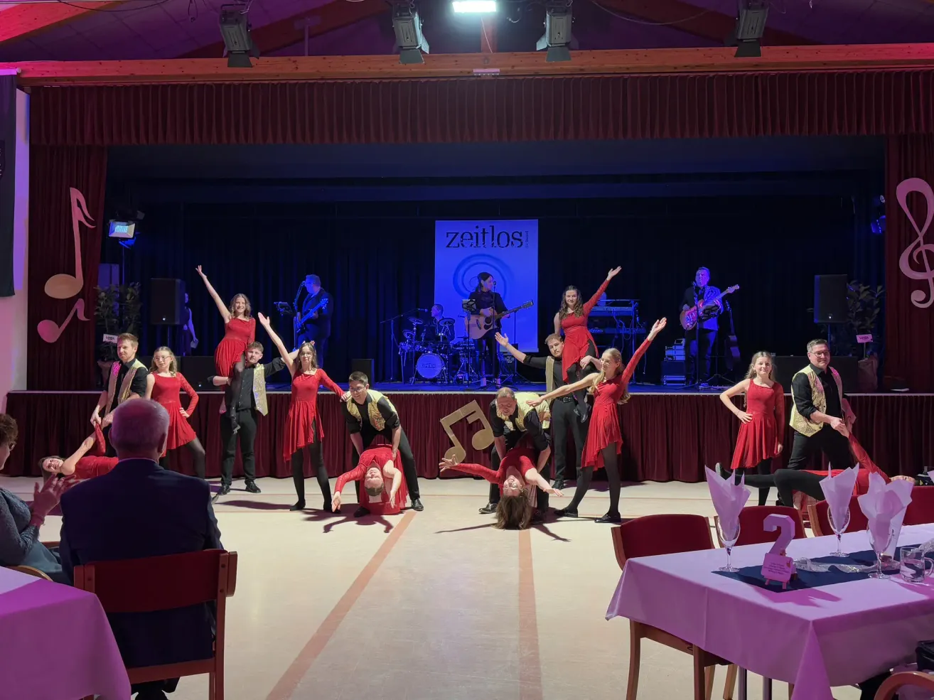 A group of dancers in red dresses perform on a stage with a sign reading 'Zeitlos'. Behind them, musicians play instruments, including guitars. The audience watches from the front row.