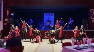 A group of dancers in red dresses perform on a stage with a sign reading 'Zeitlos'. Behind them, musicians play instruments, including guitars. The audience watches from the front row.