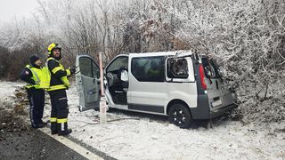 A fireman in a yellow and black jacket is standing next to a damaged white van with its back door open and a broken window on a snowy road.