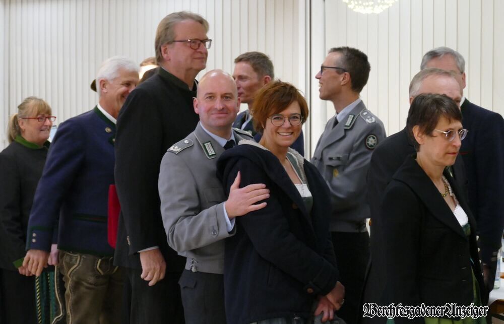 A group of people in uniforms and suits stand together in a room. A man in uniform hugs a woman smiling.