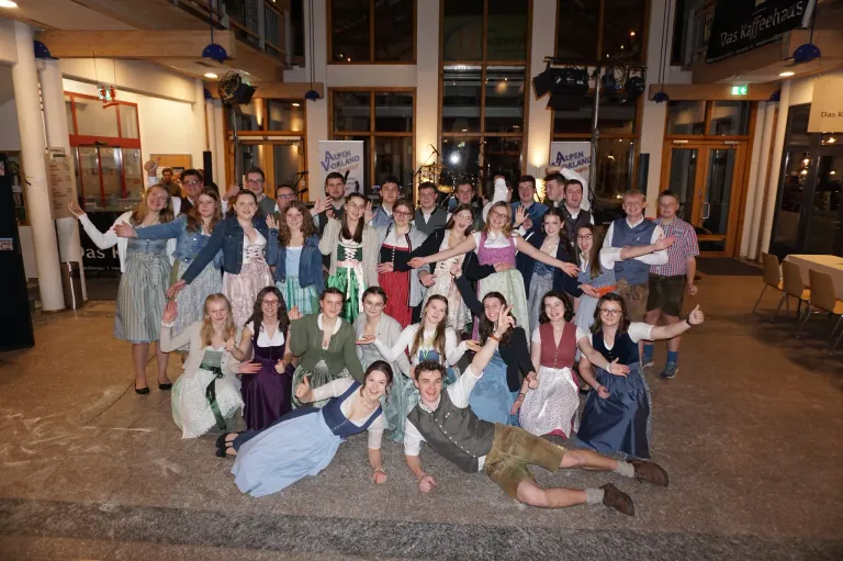 Eine Gruppe von Menschen in traditioneller Kleidung, möglicherweise für ein kulturelles Ereignis, posiert für ein Foto in einem Saal mit Glasfenstern und Lampen, die von der Decke hängen.