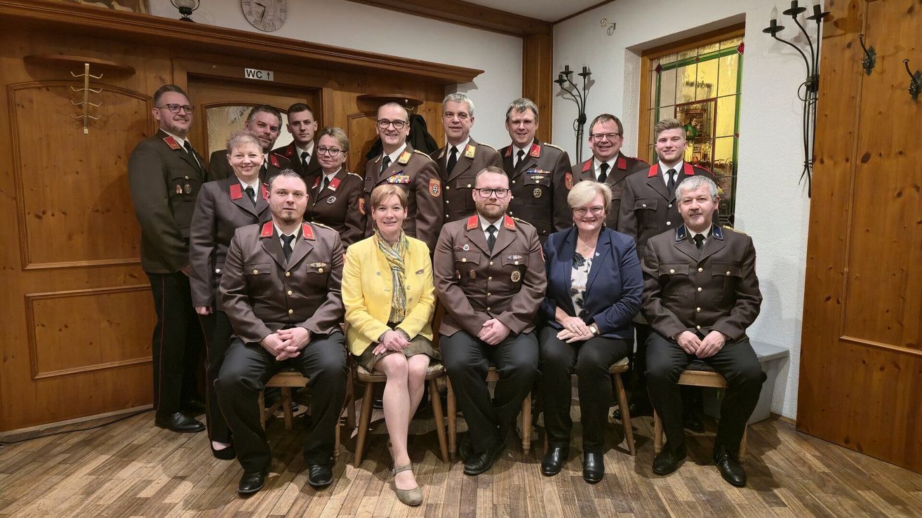 A group photo of uniformed individuals, including men and women, sitting on chairs in a room with a wooden floor. They appear to be posing for a photograph.