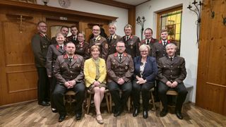 A group photo of uniformed individuals, including men and women, sitting on chairs in a room with a wooden floor. They appear to be posing for a photograph.