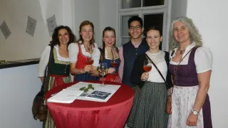 A group of people, some in traditional attire, gather around a table with wine glasses, smiling for a photo. The setting appears to be a social event, with books and papers on the table.