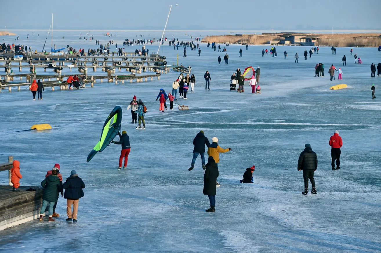 Many people skate on frozen water, some holding kites, while others sit on the ice. A few are wearing hats, gloves, and shoes. The ice surface is wide and clear, with several wooden benches nearby.