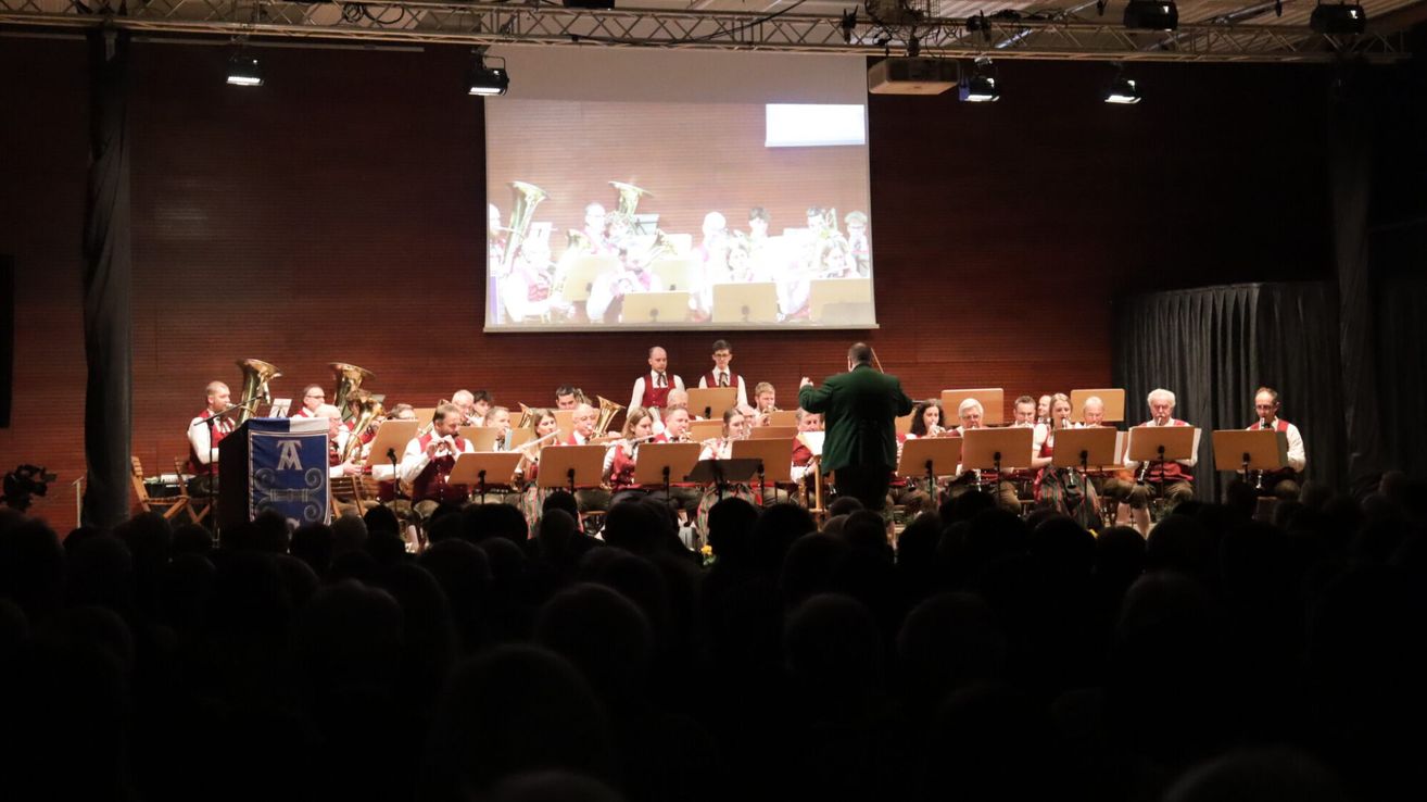 A concert with an orchestra and brass band is happening. A large screen displays the musicians. The audience is watching.