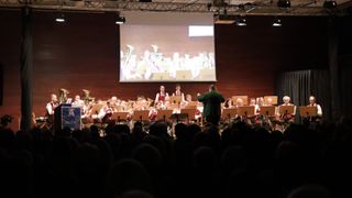 A concert with an orchestra and brass band is happening. A large screen displays the musicians. The audience is watching.