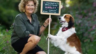 An older woman with short curly hair and glasses holds a sign with a dog standing next to her. The sign reads 'Therapie Begleithund Harley + Christa' with a date 'Okt. 2025'. The dog is wearing a red and white plaid collar.