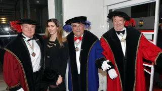Four individuals, adorned in ceremonial attire, stand together in front of a building. They wear hats, medals, and white gloves. The man on the right has a red feather on his hat.