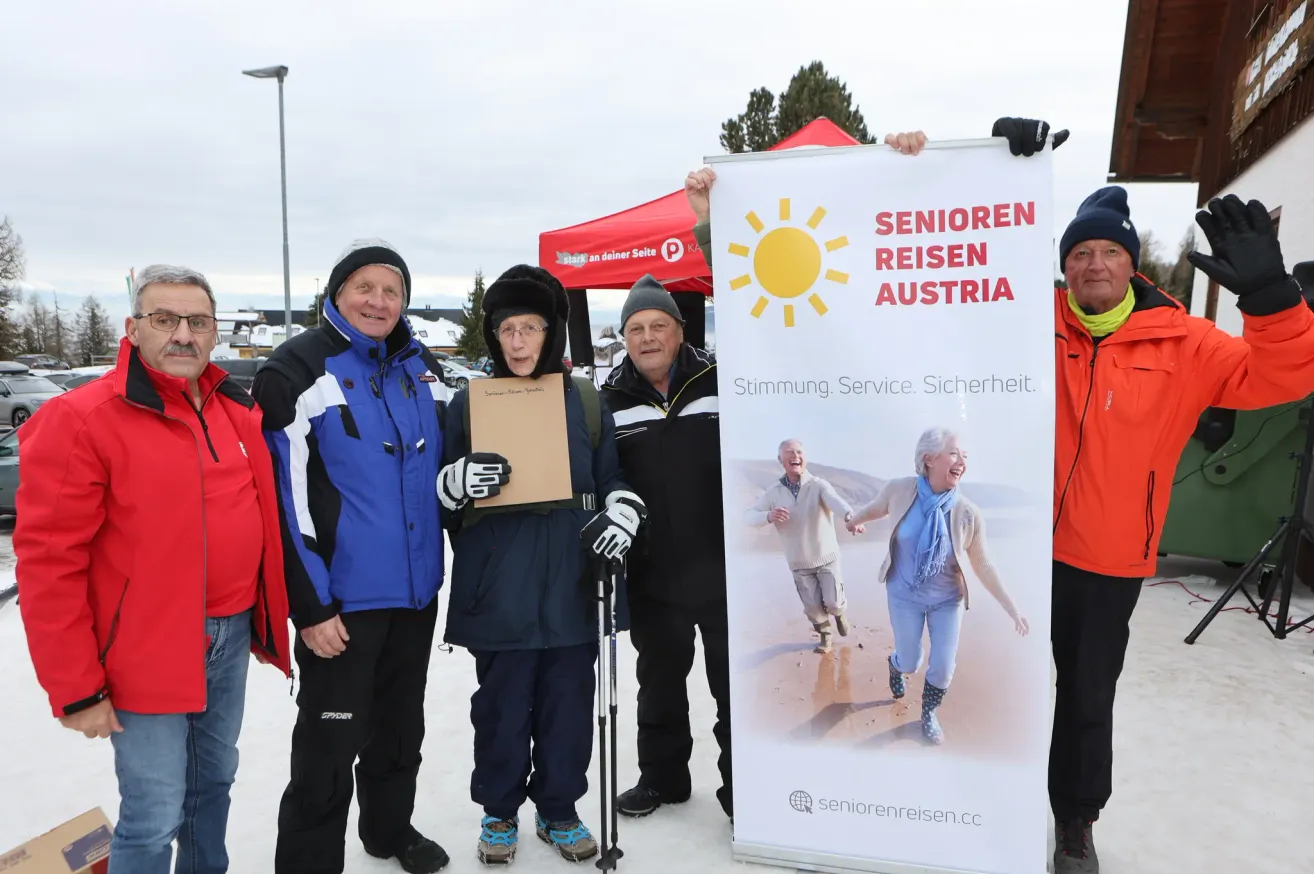 Five adults in winter clothing pose with a banner for Senioren Reisen Austria. The banner has a sun and the words Senioren Reisen Austria in red.