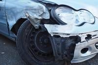 Ein blaues Auto hat Schäden am vorderen Stoßfänger und Scheinwerfer erlitten, mit sichtbaren Kratzern und Dellen an der vorderen Stoßstange und der Motorhaube.