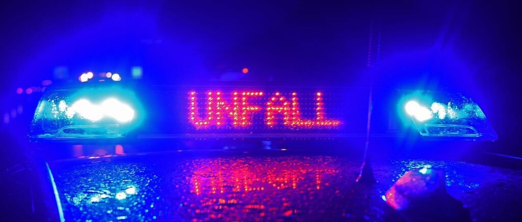 Blue and red lights flash on a wet road at night, with a sign above reading 'UNFALL', likely indicating an accident.