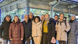 Eine Gruppe von Menschen posiert für ein Foto vor einer Baustelle mit beleuchteten Fenstern und Stahlkonstruktionen. Sie tragen Winterkleidung.