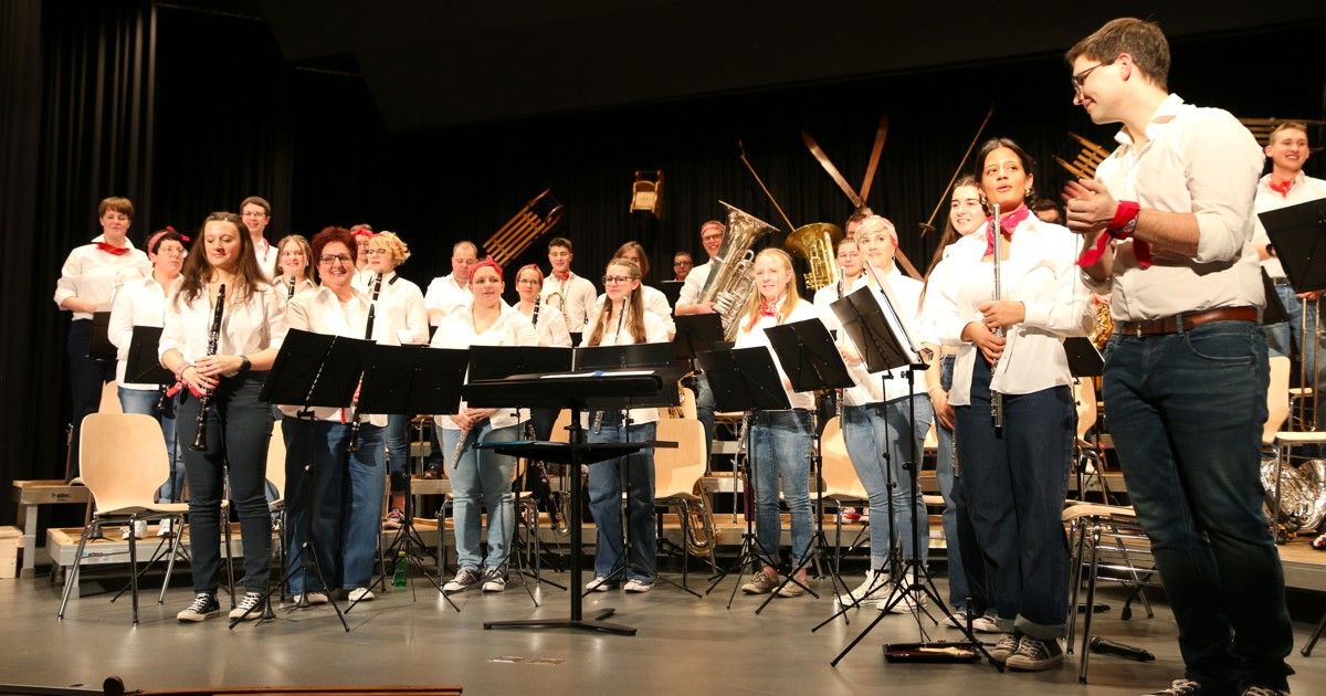 A group of musicians, including clarinetists and trumpet players, are on stage holding their instruments. They are dressed in white shirts and blue jeans, and the stage is set with music stands and black chairs.