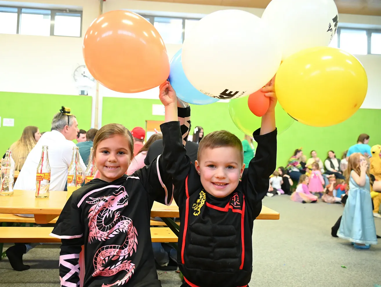 Two children in dragon themed costumes hold up balloons in a room with tables, bottles, and people sitting on the floor.