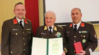 Drei Männer in Militäruniformen stehen eng beieinander. Der Mann in der Mitte hält ein Zertifikat in den Händen. Der Mann auf der linken Seite lächelt, während der Mann auf der rechten Seite eine rote Mappe in den Händen hält.