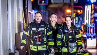 Drei Feuerwehrleute stehen lächelnd in ihren Uniformen draußen, mit einem Helm und Ausrüstung in der Hand. Die Szene ist nachts mit Straßenlaternen und einem verschwommenen Hintergrund.