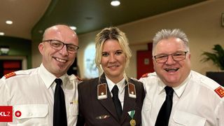 Three uniformed individuals, two men and one woman, are smiling for a photo. They all wear black ties and white shirts. The woman has a badge and a medal. Behind them, a red door is visible.
