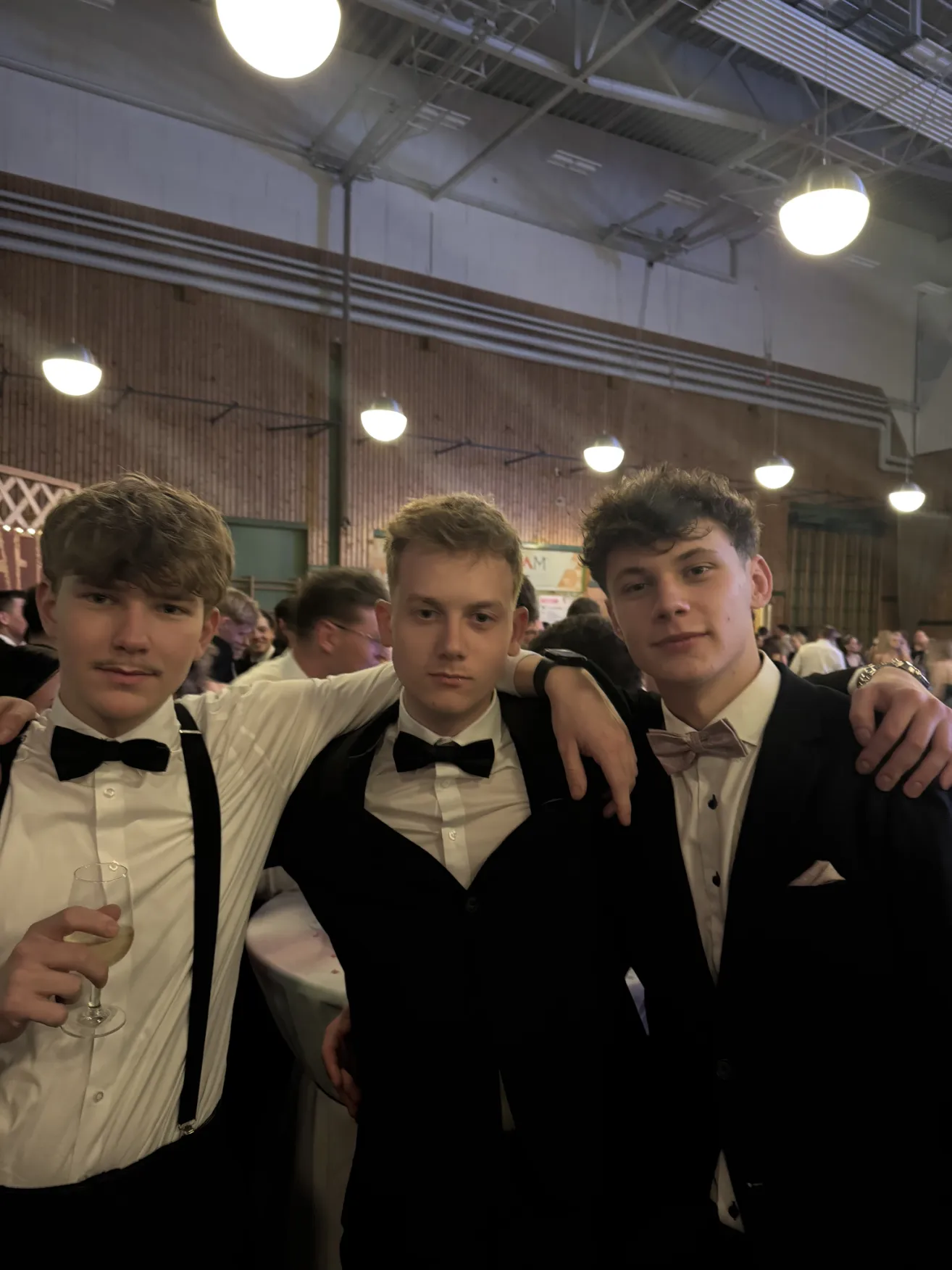 Three young men in formal attire, standing together and smiling for a photo at an event. The man on the left holds a glass of wine.
