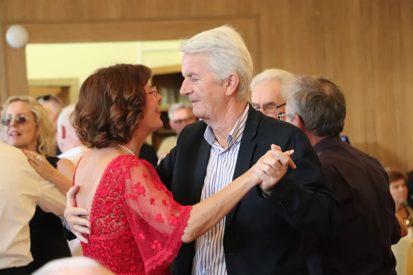 Ein älteres Paar tanzt zusammen in einem formellen Umfeld, mit einer hölzernen Wand im Hintergrund und anderen Leuten um sie herum.