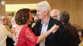Ein älteres Paar tanzt zusammen in einem formellen Umfeld, mit einer hölzernen Wand im Hintergrund und anderen Leuten um sie herum.