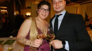 A man and a woman in formal attire are standing together, holding wine glasses filled with wine. The woman is wearing a gold scarf and has a tattoo on her left arm. They appear to be at a social event.