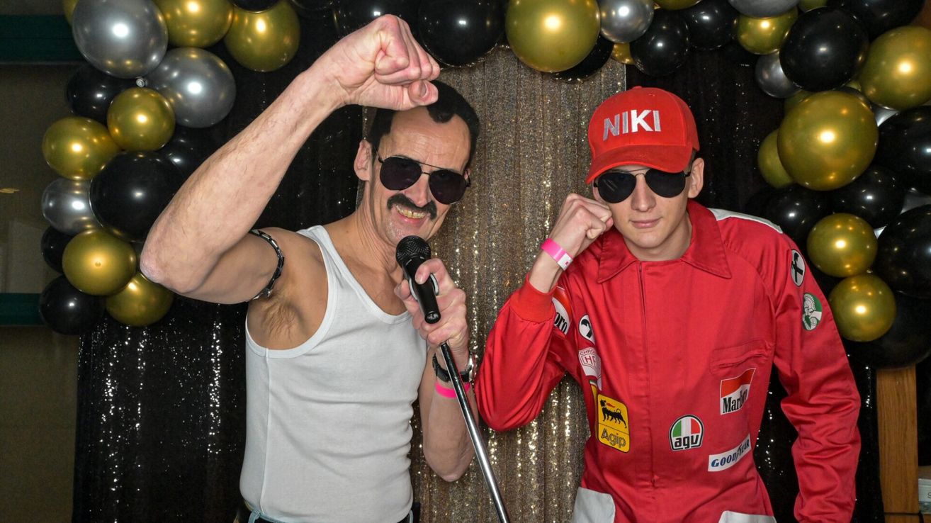 Two men in a photo booth wearing costumes, one holding a microphone, the other in a red racing suit and a red hat with the word 'NIKI' on it. Behind them is a colorful backdrop of balloons.