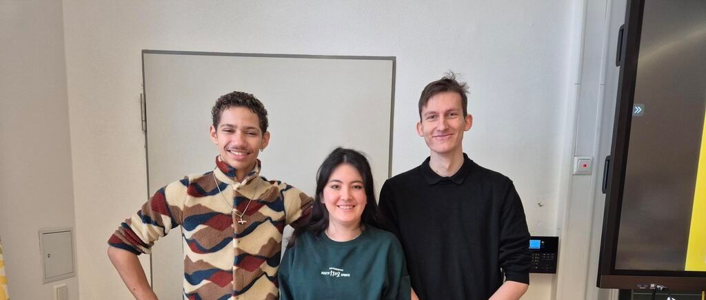 Drei Menschen stehen gemeinsam in einem Raum, lächeln und posieren für ein Foto. Ein Whiteboard ist an der Wand hinter ihnen montiert.