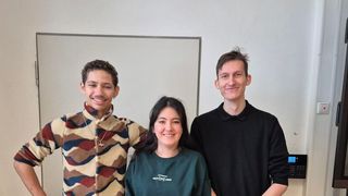 Drei Menschen stehen gemeinsam in einem Raum, lächeln und posieren für ein Foto. Ein Whiteboard ist an der Wand hinter ihnen montiert.