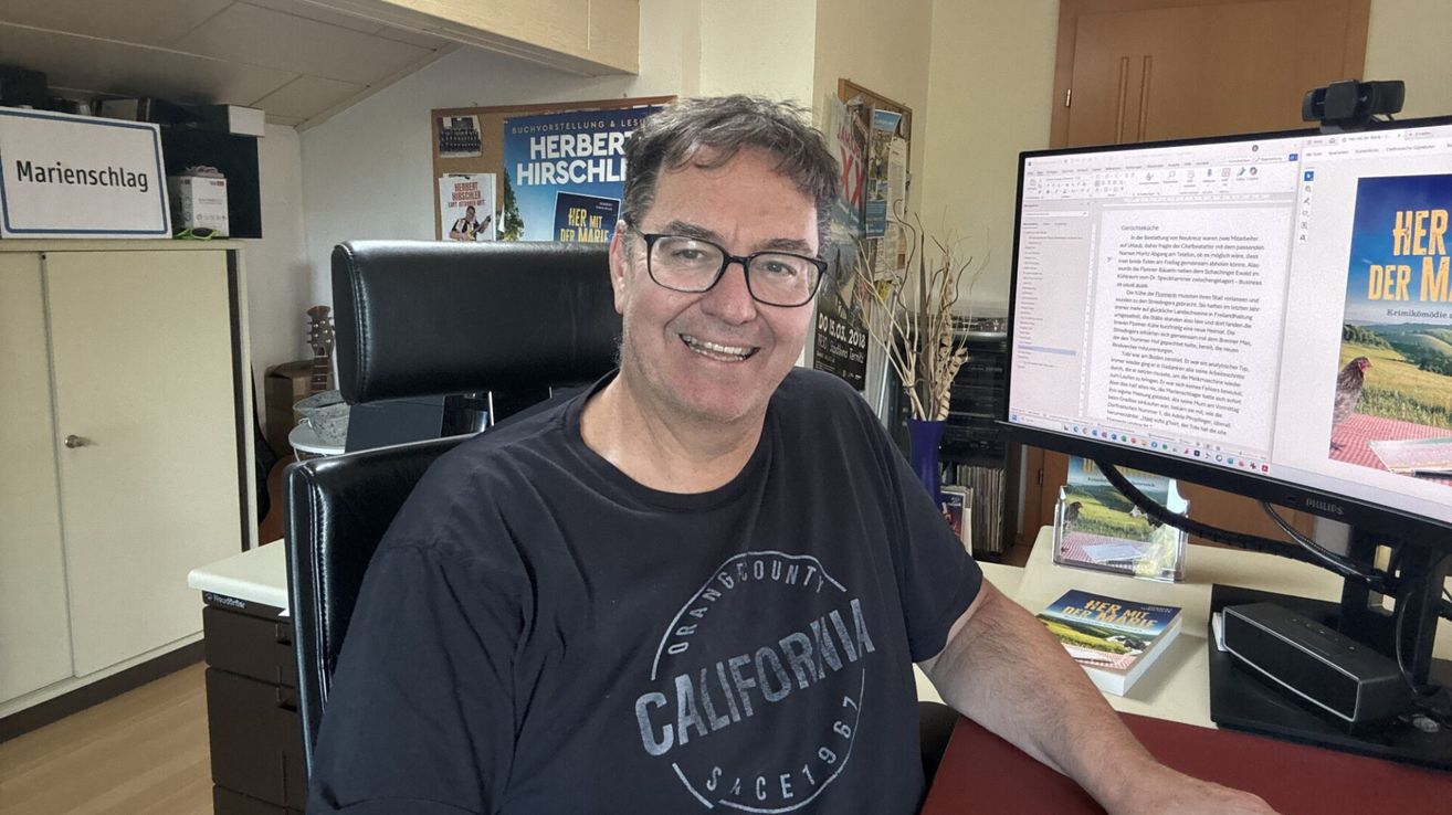 A man is seated at a desk, smiling. He wears glasses and a black t-shirt with the text 'Orange County California since 1987.' Behind him, there is a monitor, books, and a poster.