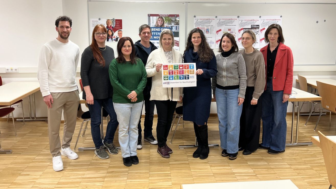 Eine Gruppe Erwachsener posiert für ein Foto mit einem Poster, das 'Dezember für Nachhaltigkeit' sagt. Sie stehen in einem Raum mit Holzböden.