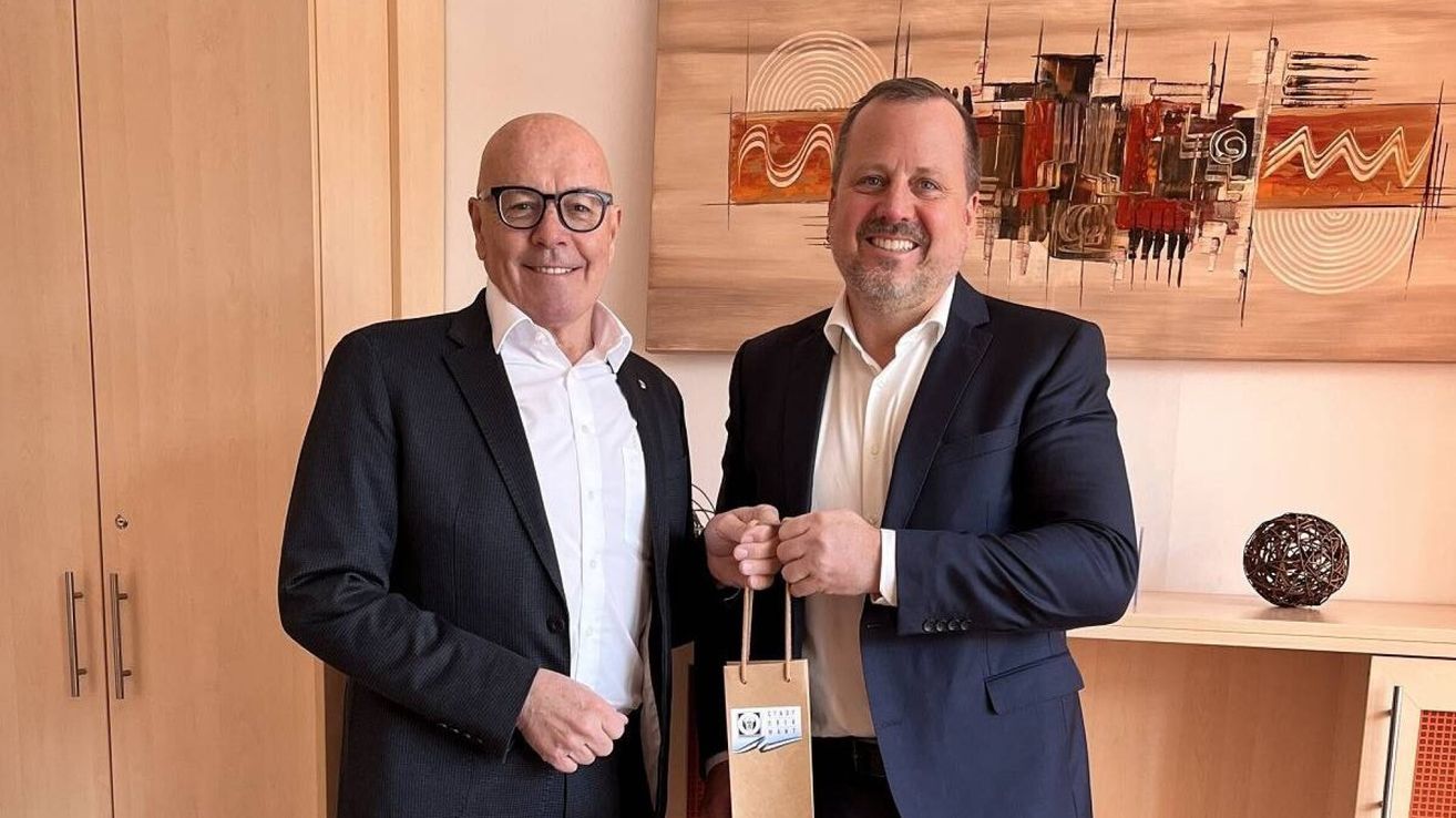 Two men in business attire stand next to each other, one holding a paper bag. They are smiling and appear to be posing for a photo. Behind them is a large, abstract painting on the wall.