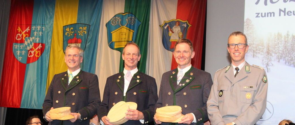 Vier Männer in traditionellen Uniformen stehen vor Flaggen. Sie halten Holzobjekte. Zwei auf der linken Seite tragen grüne Uniformen, während die beiden auf der rechten Seite Militäruniformen tragen.