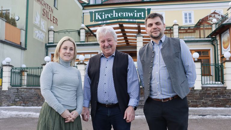 Drei Personen stehen vor einem Gebäude mit dem Schild 'Kirchenwirt'. Die Frau hat blonde Haare, der Mann in der Mitte lächelt und der Mann rechts trägt eine Weste.