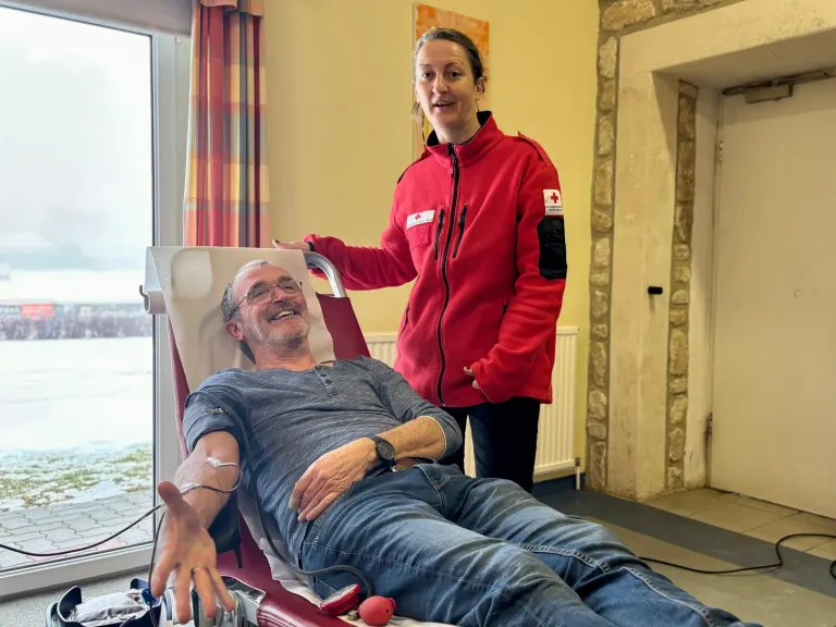 Ein lächelnder Mann liegt auf einem roten Stuhl mit einem Blutspendegerät, begleitet von einer Frau in einer roten Jacke.