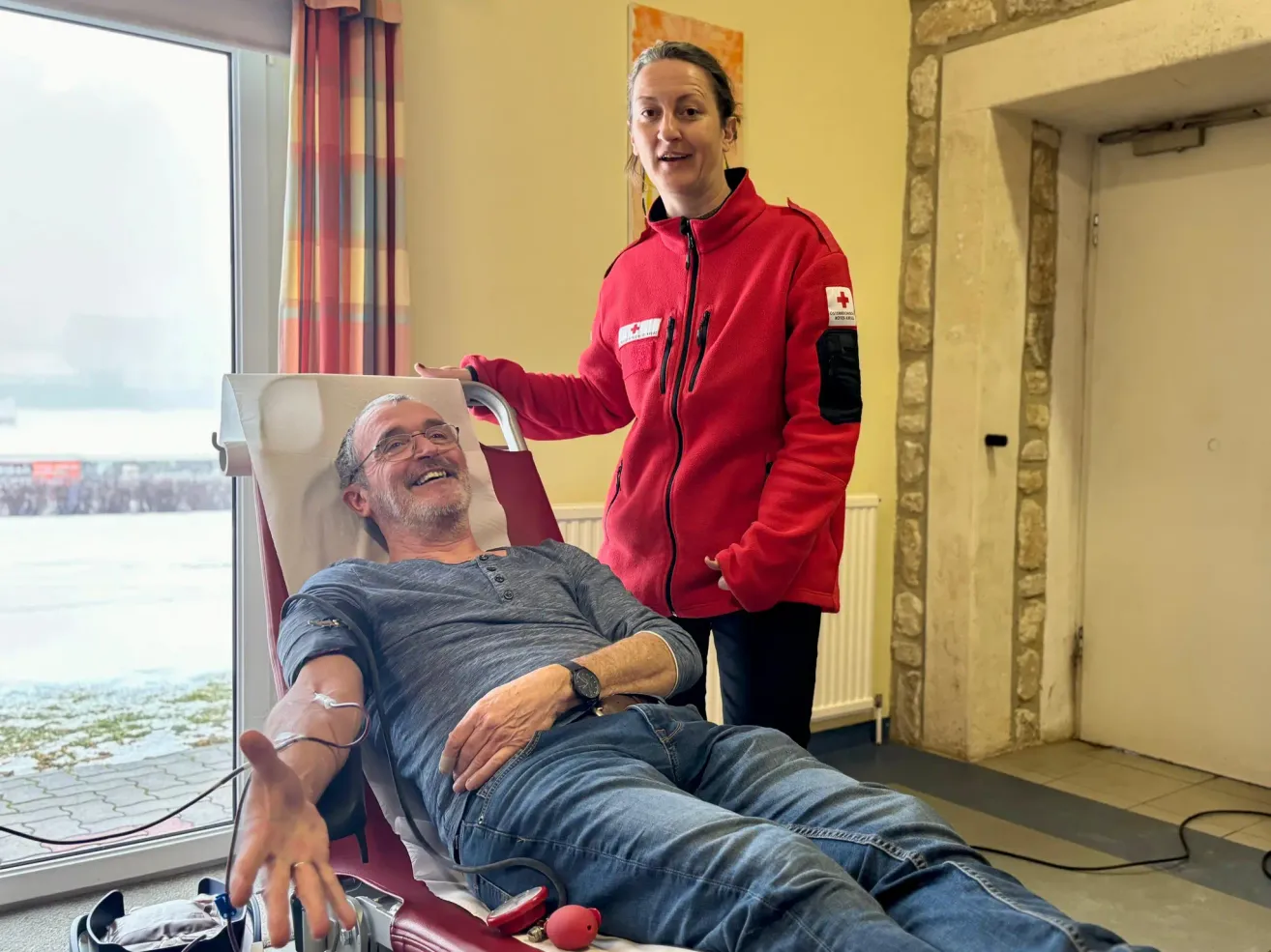Ein lächelnder Mann liegt auf einem roten Stuhl mit einem Blutspendegerät, begleitet von einer Frau in einer roten Jacke.