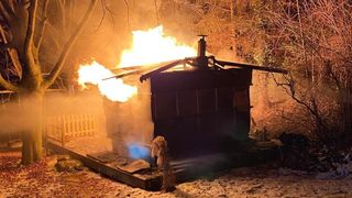 Eine Holzstruktur ist von Flammen umhüllt, mit aufsteigendem Rauch. Das Feuer ist intensiv und wirft ein warmes Licht auf die Umgebung.