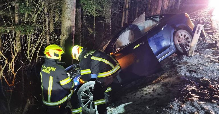 Zwei Feuerwehrleute in Schutzkleidung arbeiten an einem umgekippten blauen Auto im Wald.