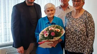 An elderly woman holding a bouquet of flowers stands between two older men and a younger man, all smiling for a photo in a room with white curtains and a white radiator.