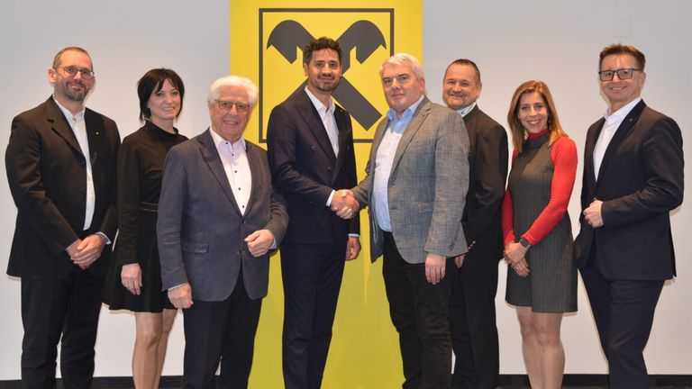 Sechs Personen in formeller Kleidung stehen vor einem gelben Hintergrund mit einem großen schwarzen Werkzeugsymbol. Zwei Männer in der Mitte schütteln sich die Hände.