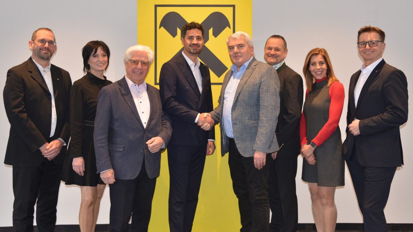 Sechs Personen in formeller Kleidung stehen vor einem gelben Hintergrund mit einem großen schwarzen Werkzeugsymbol. Zwei Männer in der Mitte schütteln sich die Hände.