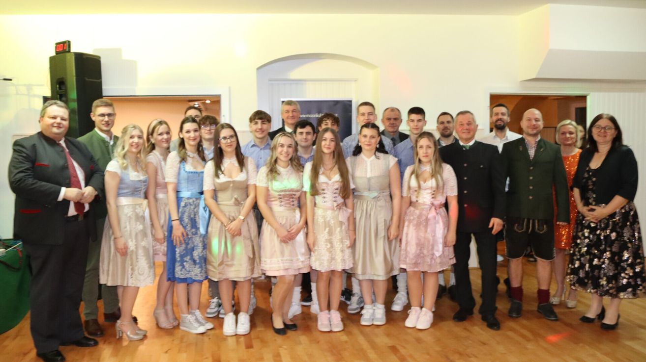 Eine Gruppe von Menschen, darunter junge Frauen in traditioneller deutscher Kleidung und Männer in Anzügen, posiert für ein Foto in einem Raum mit Holzböden und einer weißen Wand.