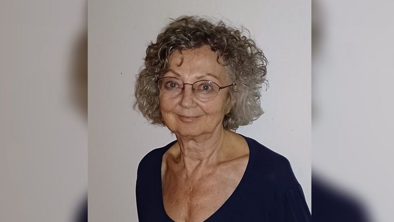 Eine ältere Frau mit grauen lockigen Haaren, die eine Brille trägt und ein marineblaues V-Neck-Shirt anhat, lächelt vor einer weißen Wand.