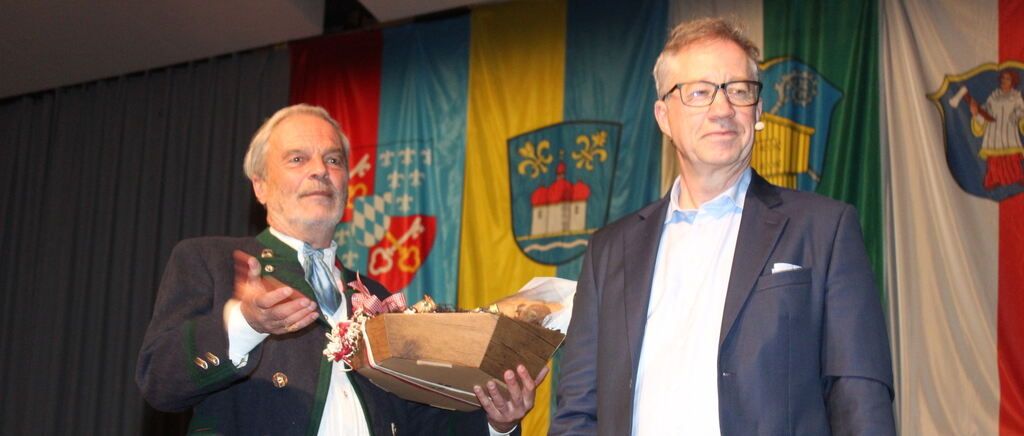 Two men stand in front of a decorated banner, one wearing a traditional outfit and holding a box, the other in formal attire with a microphone.
