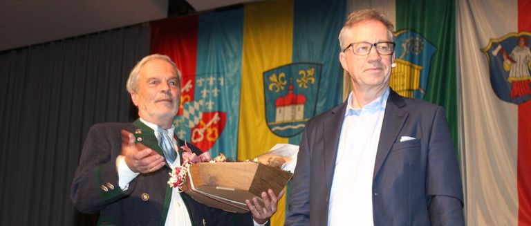 Two men stand in front of a decorated banner, one wearing a traditional outfit and holding a box, the other in formal attire with a microphone.