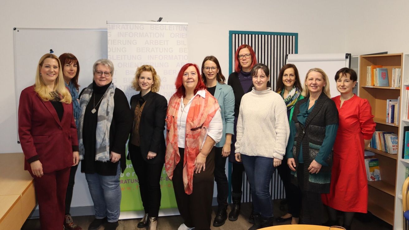 Eine Gruppe von Frauen, alle formell gekleidet, steht in einer Büroumgebung zusammen. Die Frau in der Mitte trägt ein rotes Kopftuch und ein weißes Hemd, während andere Jacken und Brillen tragen. Sie stehen vor einem Whiteboard mit deutschem Text.