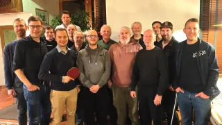 A group of men stands together in a room with a white backdrop. One man holds a ping pong paddle. They are dressed in casual clothing, some wearing glasses and hats.