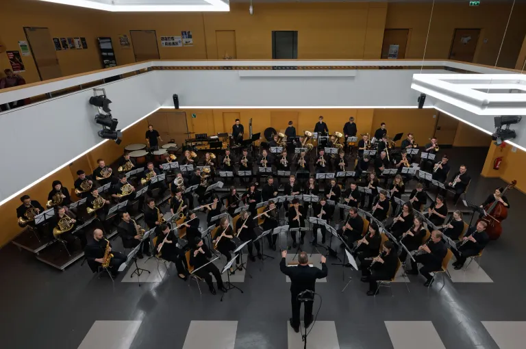A large orchestra performs in a hall, with musicians playing various instruments. The conductor stands in the center, leading the ensemble. The audience is seated in chairs around the orchestra.