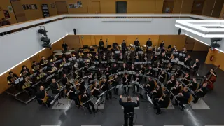 A large orchestra performs in a hall, with musicians playing various instruments. The conductor stands in the center, leading the ensemble. The audience is seated in chairs around the orchestra.
