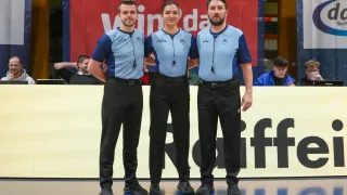 Three referees stand on a blue floor, dressed in matching uniforms with belts. The woman in the middle has a smile on her face. Behind them is a yellow banner with red text. Two people are sitting behind the banner.
